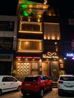 Un hotel con un cartel que dice 'Hotel Mohan Tulip' y un cartel que anuncia 'Habitaciones | Restaurante | Salón de banquetes'. Hay dos coches estacionados al frente.