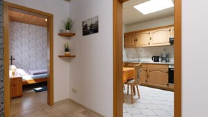 A hallway leading to a bedroom with a bed, a small bedside table, and a nightstand. There are two wooden shelves with plants on the wall. The room has a tiled floor and a window with a view of buildings. - Bungalow Fischerstraße 6 (Wittmund)