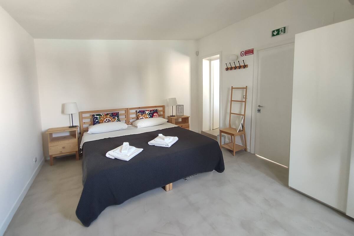 Une chambre avec un grand lit, deux tables de chevet, une chaise, un porte-manteau et une porte portant un panneau indiquant la sortie de secours.