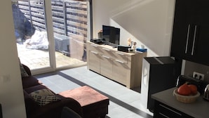 Eine moderne Küche mit Essbereich, einer Couch und einer Schiebetür zu einer Terrasse mit Blick auf ein Gebäude und einen Zaun.