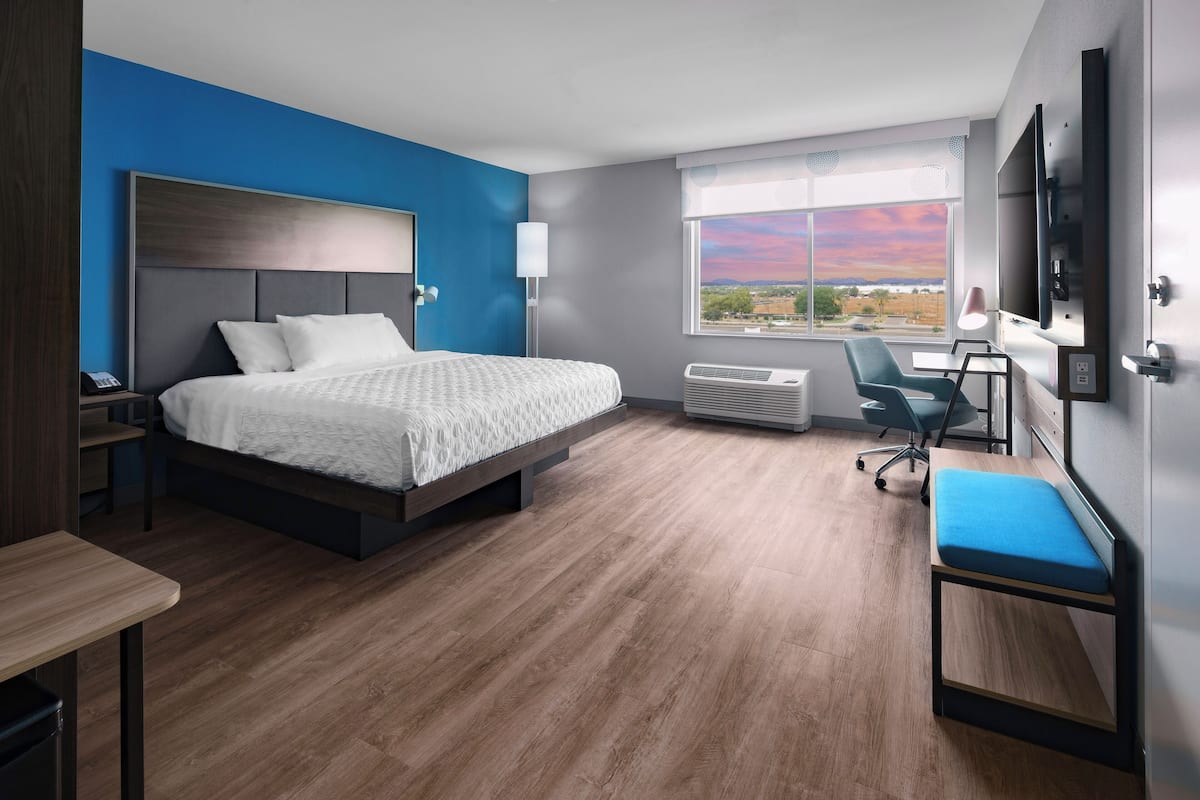 A modern hotel room with a large bed, a desk, a chair, a TV, and a window with a view.