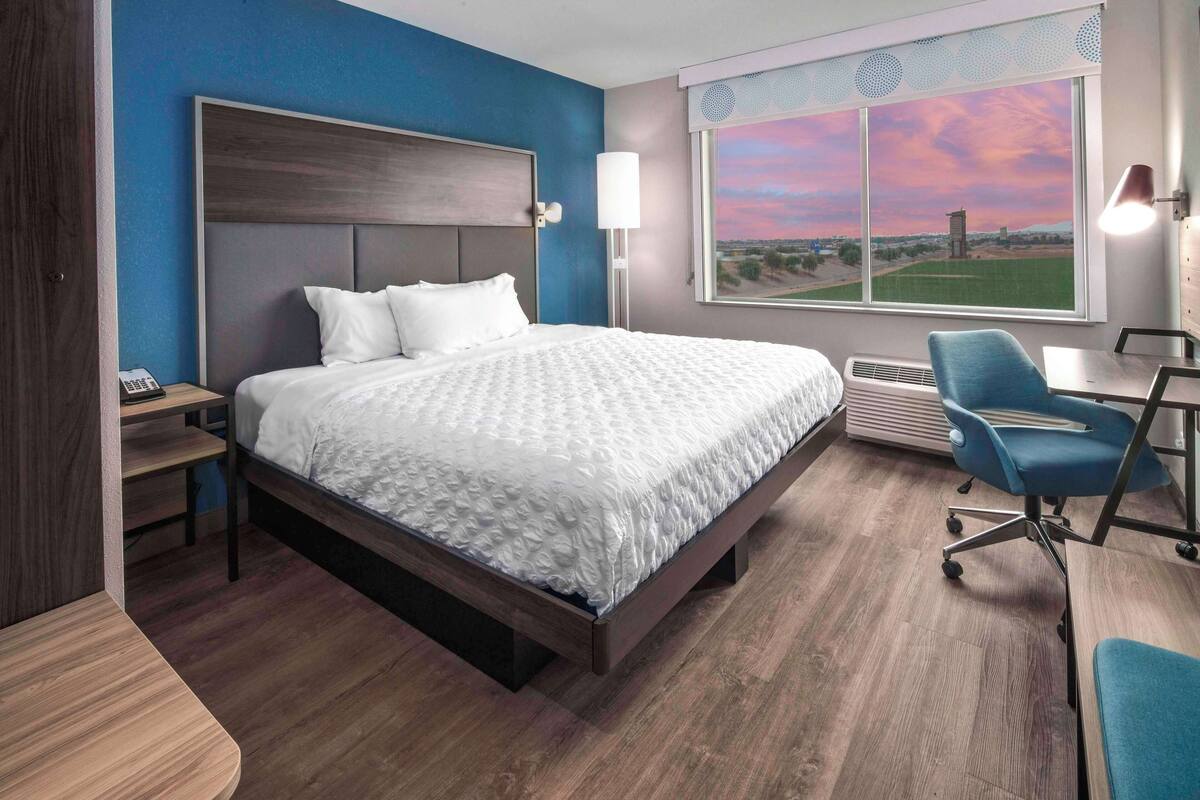 A hotel room with a large bed, a desk with a chair, a window with a view, and a nightstand with a phone.