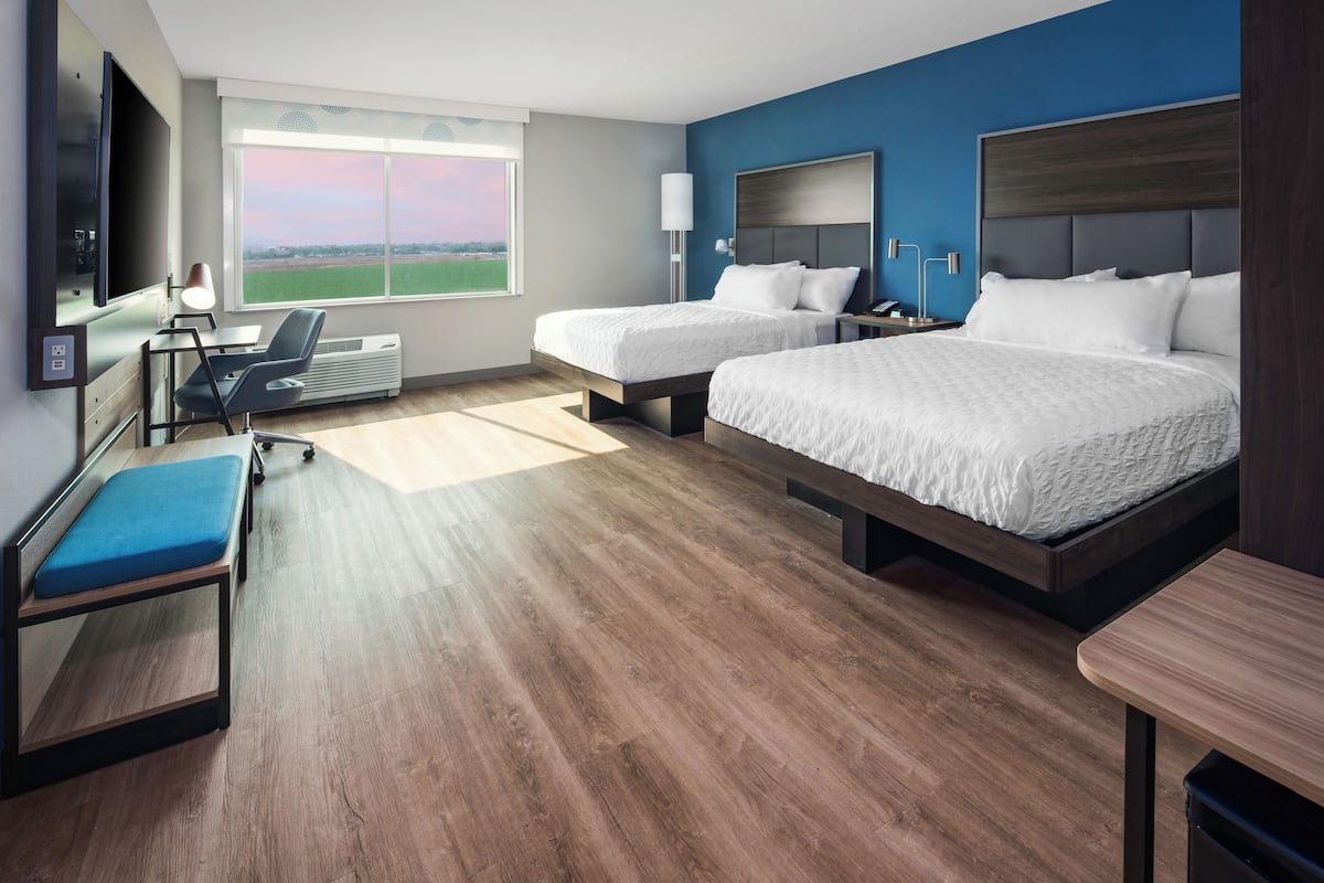 A hotel room with two beds, a desk, a chair, a TV, and a large window with a view of a field.