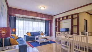 Una sala de estar con un sofá, una mesa de centro y sillas. Hay un televisor y una estantería.