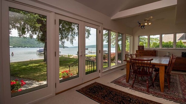 A spacious dining area with a large table and chairs, large windows with a view of a lake and dock, and a ceiling fan.