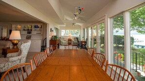 A spacious dining area with a large wooden table and chairs, a chandelier, and large windows offering a view of the outdoors.