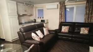 A living room with a brown leather sofa, a glass coffee table, and a black leather sofa with floral cushions. There is a kitchen area with white cabinets and a microwave. - Hiša Kramer - Apartment Borjana (Kobarid)