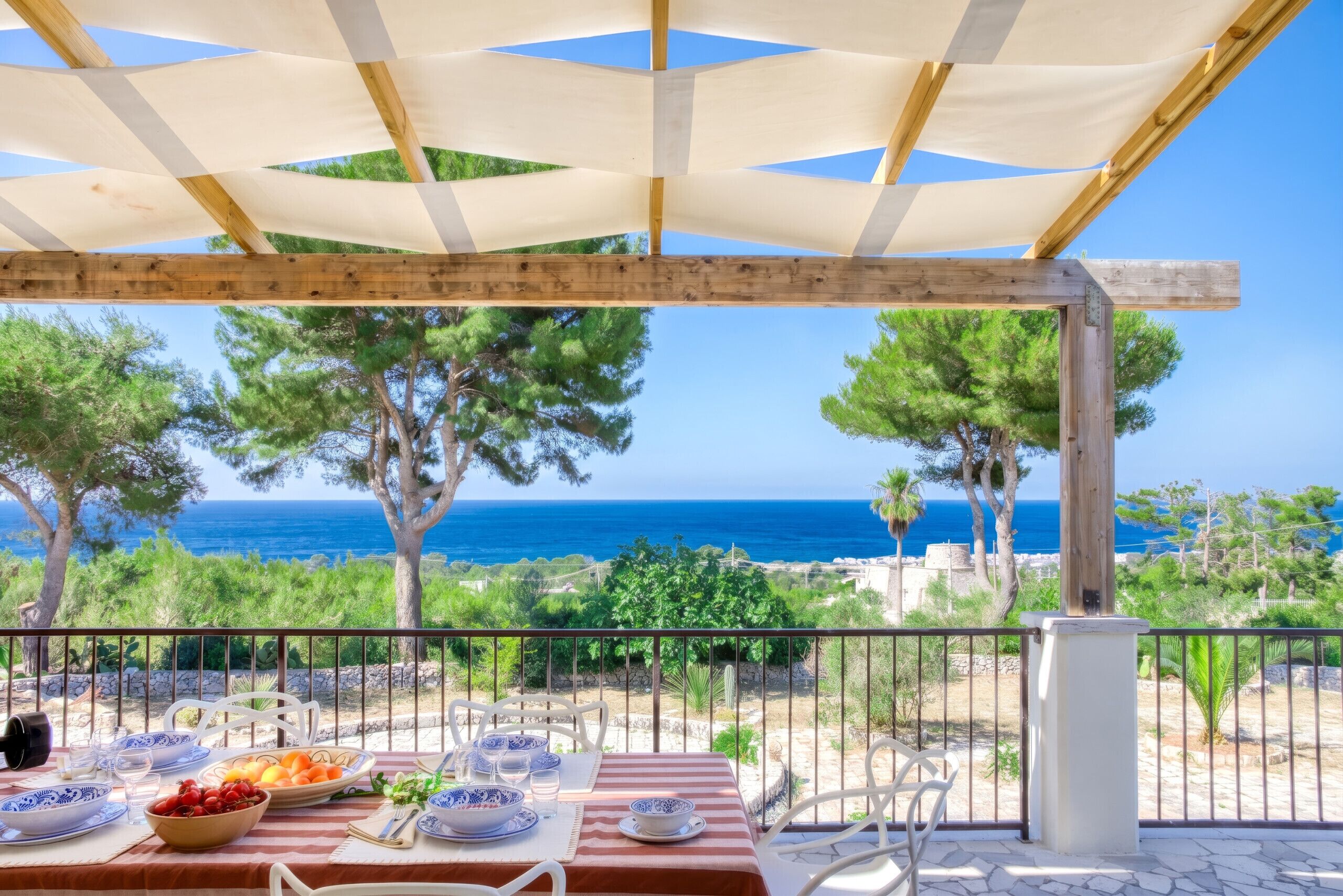 A terrace with a table set for dining, overlooking a coastal landscape with trees and the sea.
