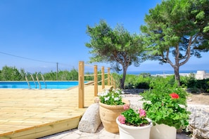 A wooden deck leading to a pool with potted plants and trees in the background.