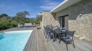 Un espace aménagé au bord de la piscine, avec une terrasse en bois, des sièges d’extérieur et un bâtiment en pierre.