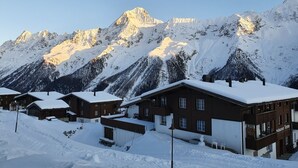 A snow-covered mountain village with chalet-style buildings, a restaurant named "Restaurant Tenne," and a clear sky.