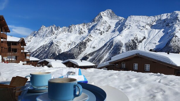 A snow-covered mountain landscape with wooden chalets and a table with cups and biscuits.