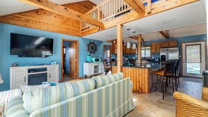 A spacious living area with a large TV, wooden ceiling beams, a striped sofa, and a kitchen with wooden cabinets and a bar area. - The Swan Shack - 4x4 Carova Beach, Pool, Hot Tub, Theater, Pets OK, Private Setting (Carova Beach)