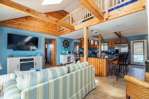 A spacious living area with a large TV, wooden ceiling beams, a striped sofa, and a kitchen with wooden cabinets and a bar area.