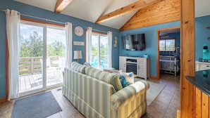 A living room with a sofa, a TV, and a sliding glass door leading to an outdoor area. - The Swan Shack - 4x4 Carova Beach, Pool, Hot Tub, Theater, Pets OK, Private Setting (Carova Beach)