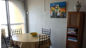 A dining area with a round table, chairs, and a bowl. There is a bookshelf with books and a painting on the wall. - Beach Front Apartment (Leucate)