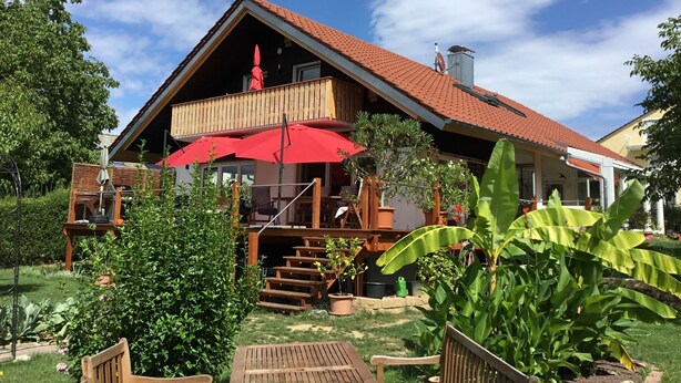 Ein zweistöckiges Holzhaus mit rotem Ziegeldach, einem Balkon mit roten Sonnenschirmen und einem Garten mit verschiedenen Pflanzen und einer Holzbank.