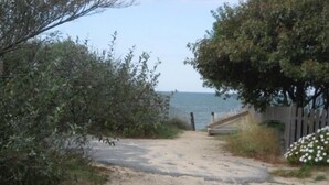Un sentiero sabbioso che conduce a una spiaggia, con una recinzione in legno e un albero a destra.