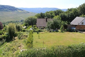 Et landhus med skrå tag, omgivet af frodig vegetation og en vingård i baggrunden.