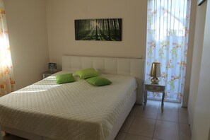 Uma cama bem arrumada com colcha e almofadas verdes, uma mesinha de cabeceira com abajur e um quadro de uma floresta na parede.