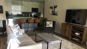 Ein modernes Wohnzimmer mit einer Couch, Couchtisch und TV-Möbel. Im Hintergrund ist eine Küche mit Mikrowelle und Barhocker zu sehen.