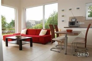 Relax in this serene space with a sofa and table near a window overlooking lush greenery, featuring a kitchen area with a microwave and a bowl.