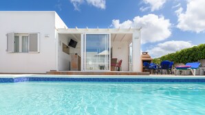 A modern poolside area with a clear blue pool, a covered seating area with blue chairs, and a white building with a sliding glass door. - Private villa with pool and whirlpool in a quiet location (Puerto del Carmen)