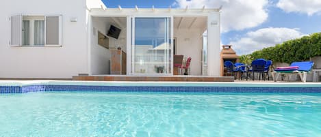 Ein modernes Haus mit Swimmingpool, einer Terrasse mit Stühlen und einem an der Wand montierten Fernseher.