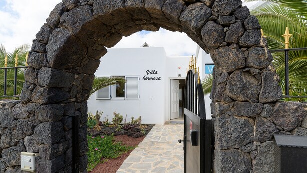 Un arc en pierre mène à un bâtiment portant l’inscription « Villa Hermosa », avec un chemin pavé et un petit espace aménagé en jardin.