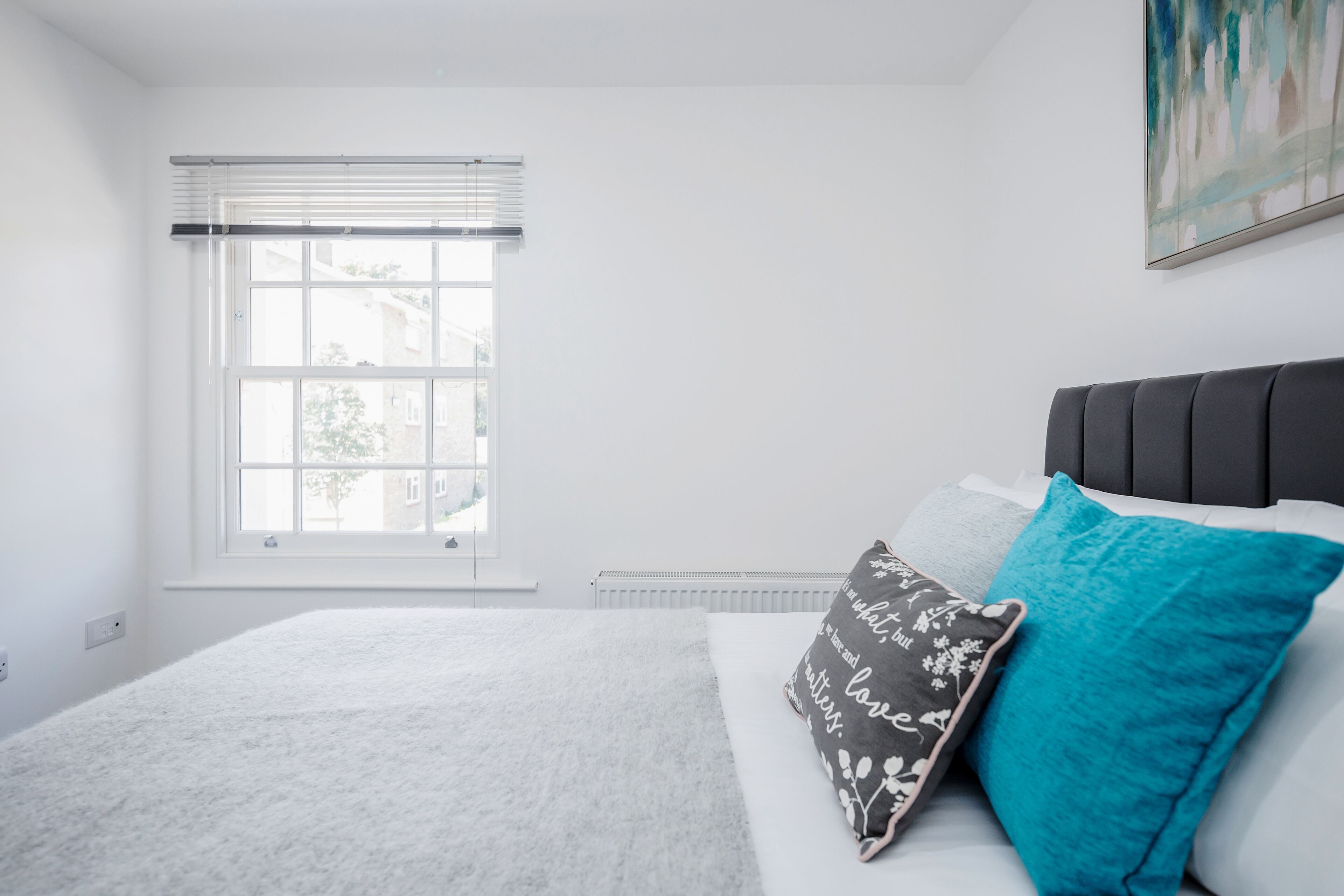 A bedroom with a bed, a window with blinds, a radiator, and a painting on the wall.