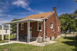 Une maison en briques avec un porche couvert, une porte d’entrée et un petit climatiseur installé au sol.