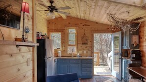 A cozy cabin interior with a wooden ceiling, a ceiling fan, a kitchen with blue cabinets, a dining area with a table and chairs, and a large window offering a view of the outdoors. - Fun Treehouse Getaway • Firepit • Lake Access (Goreville)