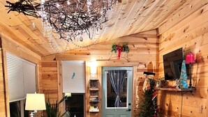 A cozy cabin interior with a dining table, a patterned armchair, a small Christmas tree, and a ceiling light fixture. - Fun Treehouse Getaway • Firepit • Lake Access (Goreville)