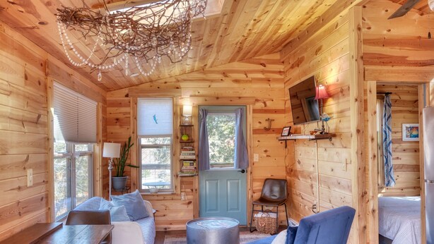 A cozy cabin interior with wooden walls, a dining area, a seating area with a blue chair, a door with a blue frame, and a ceiling with a branch chandelier. - Fun Treehouse Getaway • Firepit • Lake Access (Goreville)