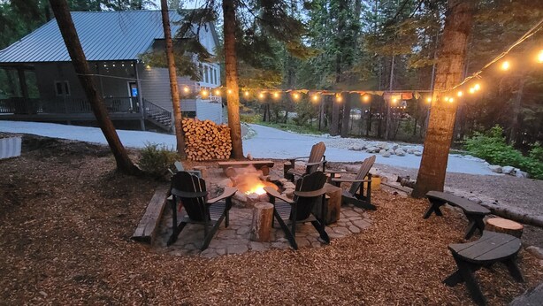 An outdoor seating area with wooden Adirondack chairs and a stone fire pit, surrounded by a wooden deck and string lights. - Luxe family and pet friendly chalet in private, forested setting (Leavenworth)