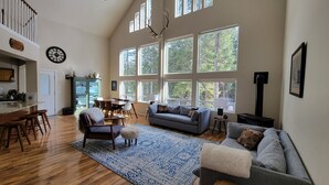 A spacious living room with a high ceiling, large windows, a dining area, and a kitchen with a green cabinet. - Luxe family and pet friendly chalet in private, forested setting (Leavenworth)