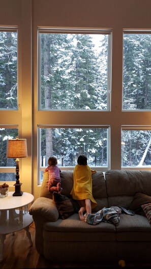 Relax in this inviting living room with a large window offering a forest view, a comfortable couch, and a small table with a lamp.