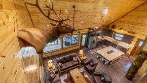 A log cabin interior with a mounted elk head, a living area with a sofa and coffee table, and a kitchen with wooden cabinets and a large window.