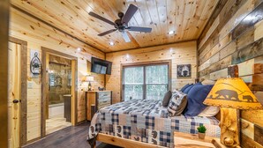 A wooden cabin bedroom with a bed, a ceiling fan, a TV, and a window with blinds.