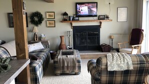 A cozy living room with a fireplace, plaid sofas, a wooden coffee table, and a television mounted above the fireplace. - Lake Ontario: Henderson, NY (Henderson)