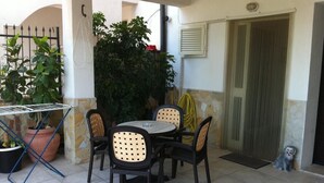 Um pátio externo com mesa redonda e quatro cadeiras, cercado por vasos de plantas e uma grande árvore.