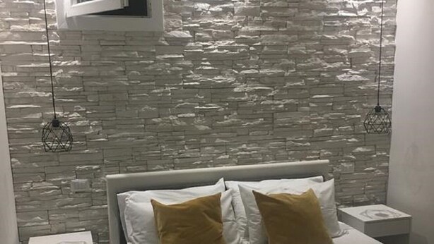 Quarto com parede de pedra, cama com roupa de cama branca e duas almofadas marrons, duas mesas de cabeceira e duas luminárias suspensas.