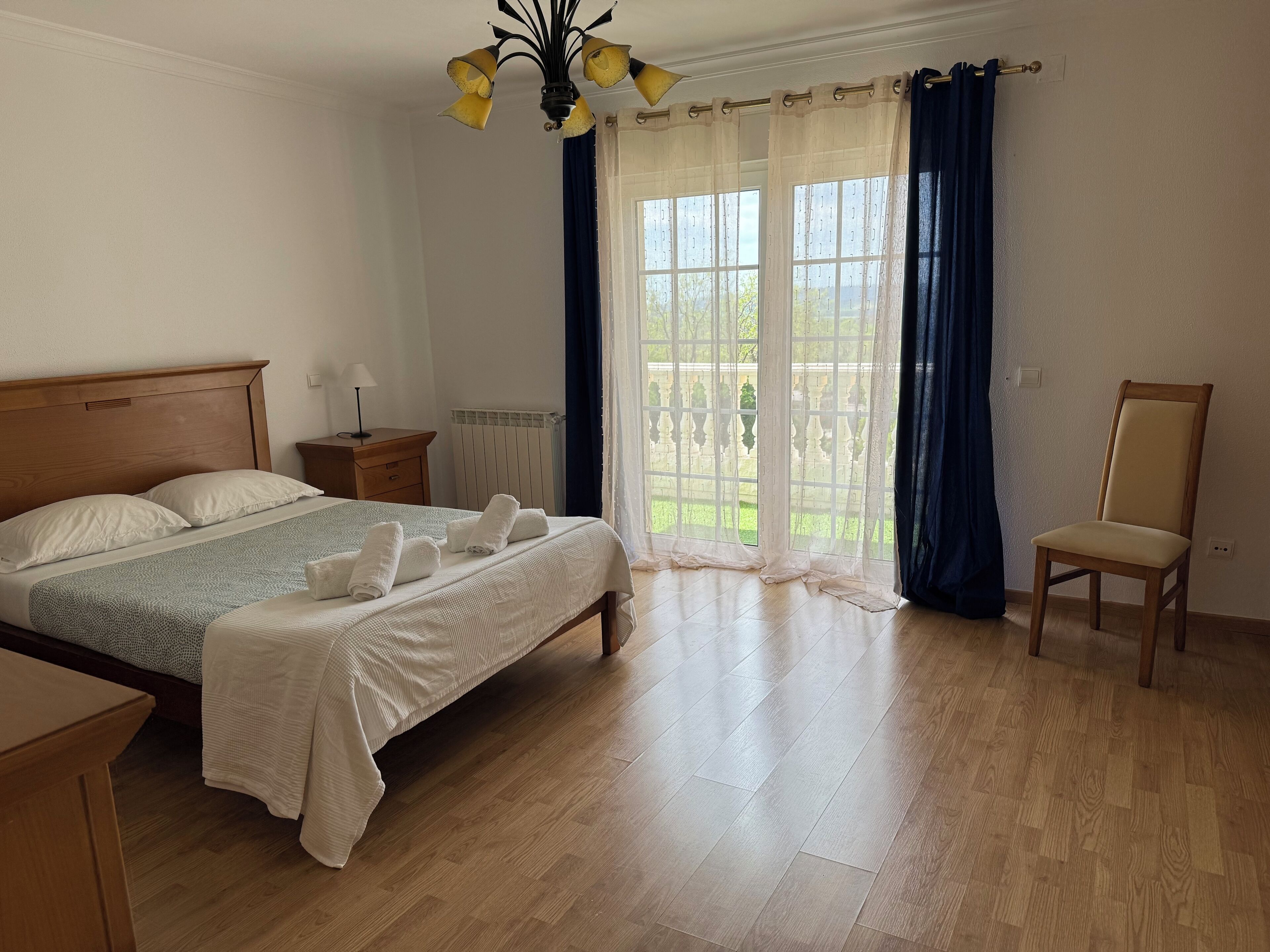 Une chambre à coucher avec un lit en bois, une table de chevet, une chaise et une vue sur l’extérieur grâce à de grandes fenêtres.