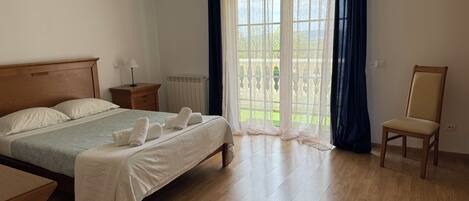 Une chambre à coucher avec un lit en bois, une table de chevet, une chaise et une vue sur l’extérieur grâce à de grandes fenêtres.