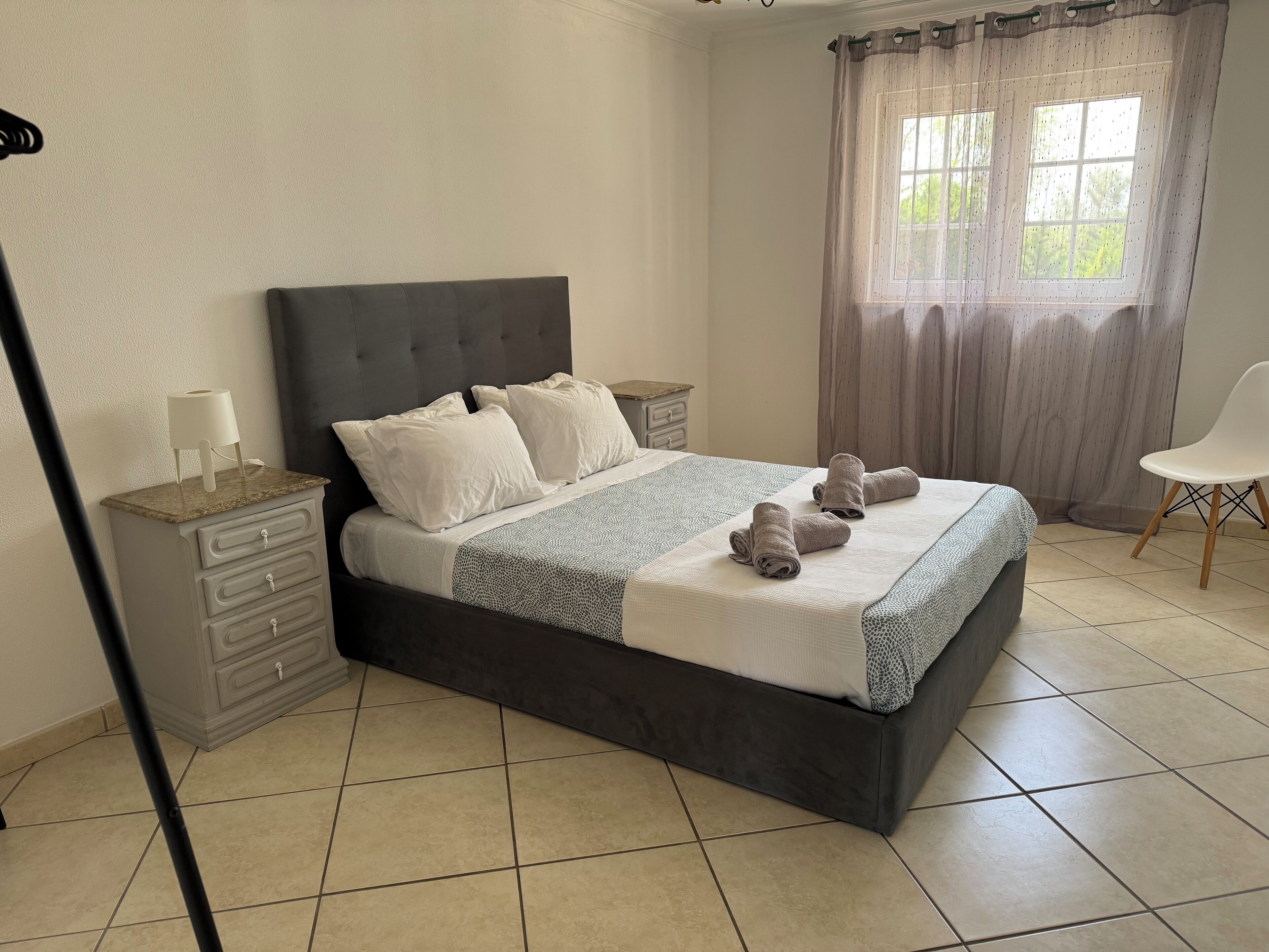 Una cama bien hecha con almohadas y una manta, una mesita de noche con lámpara, una silla y una ventana con cortinas.