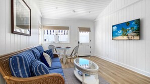 Ein Zimmer mit einem blauen, gestreiften Sofa, Korbstühlen, einem Glastisch und einem Strandbild.