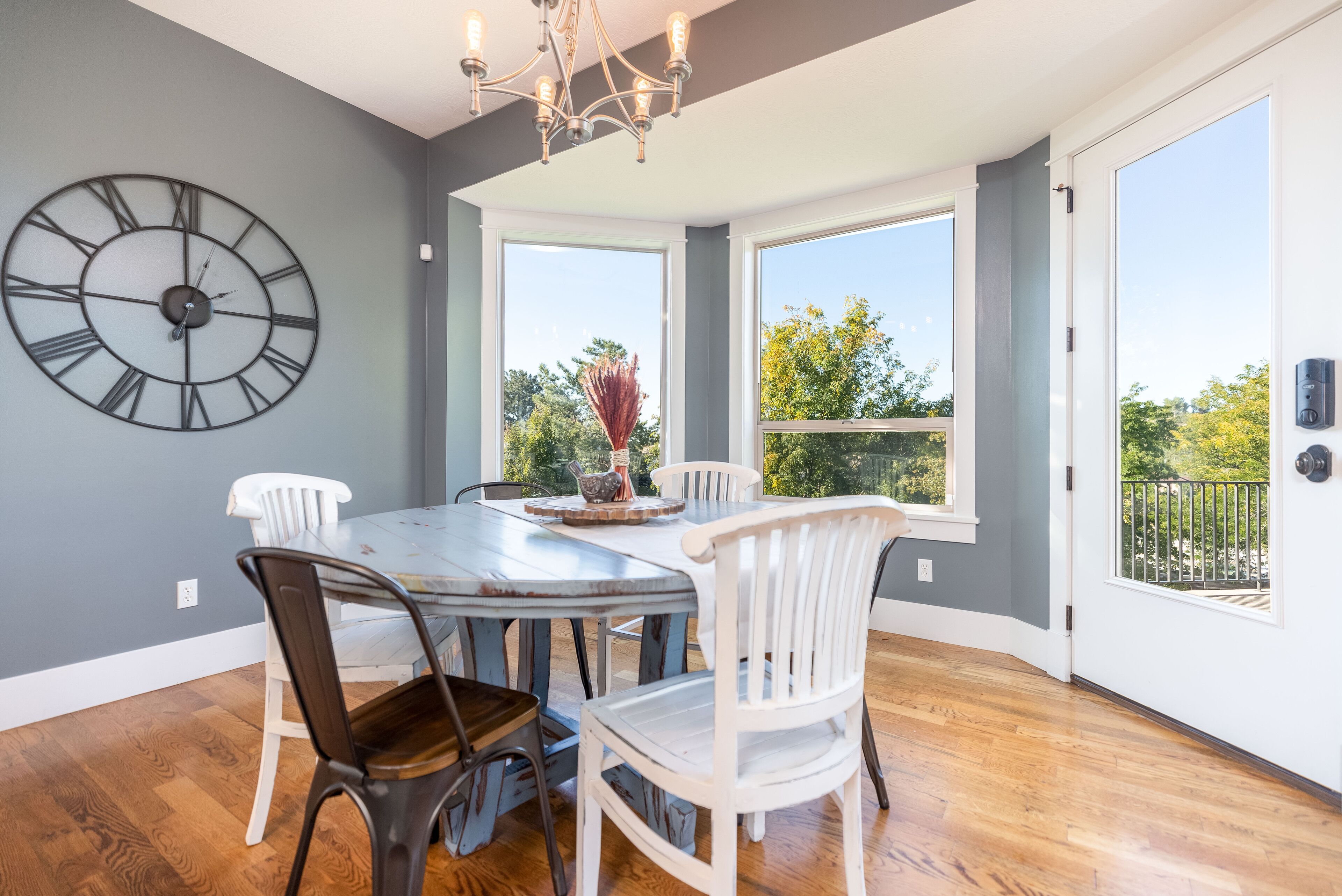 A dining area with a large wall clock, white chairs, a wooden table, and large windows offering a view of greenery.