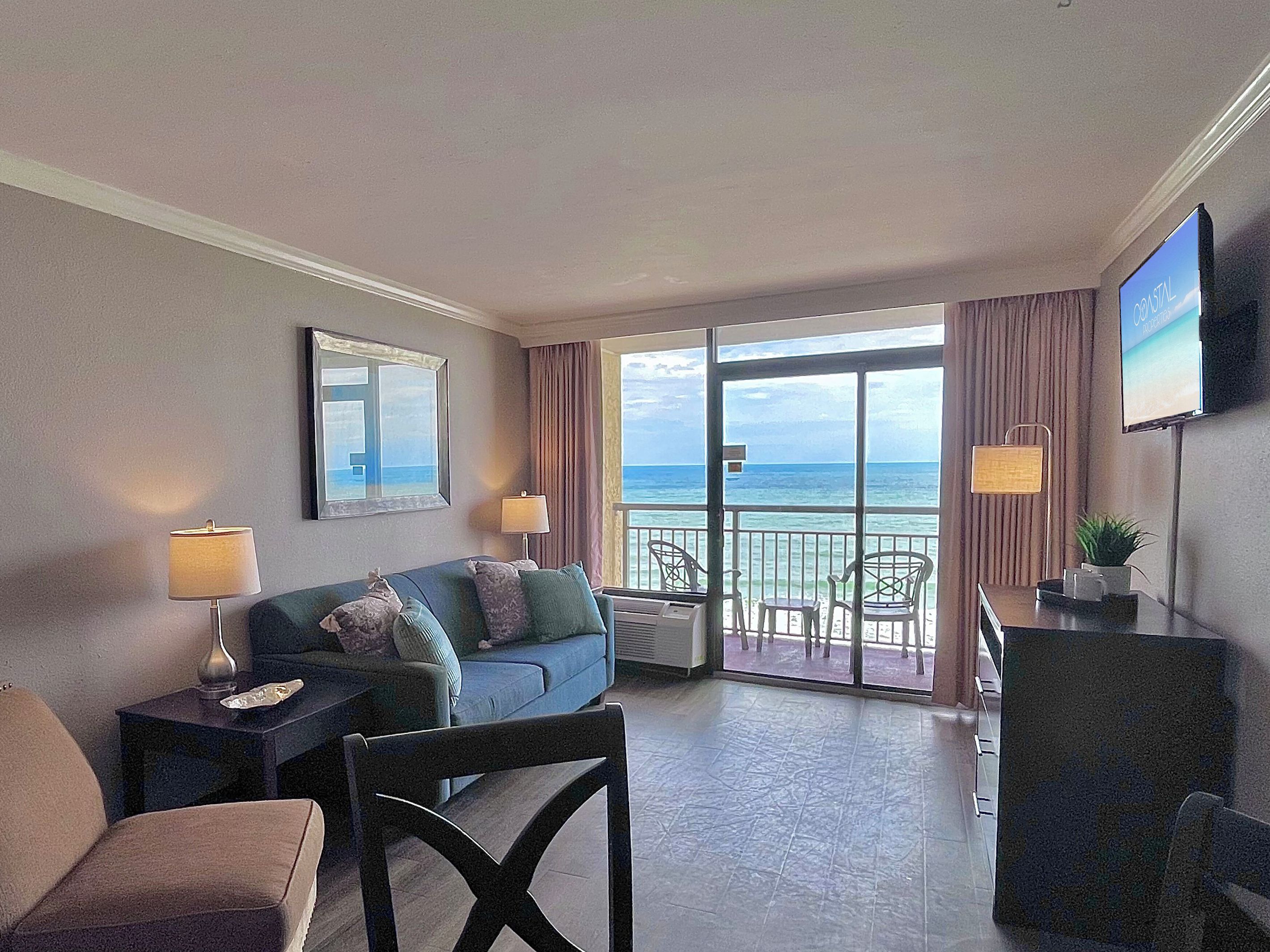 A hotel room with a balcony, a sofa, a chair, a TV, and a view of the ocean.
