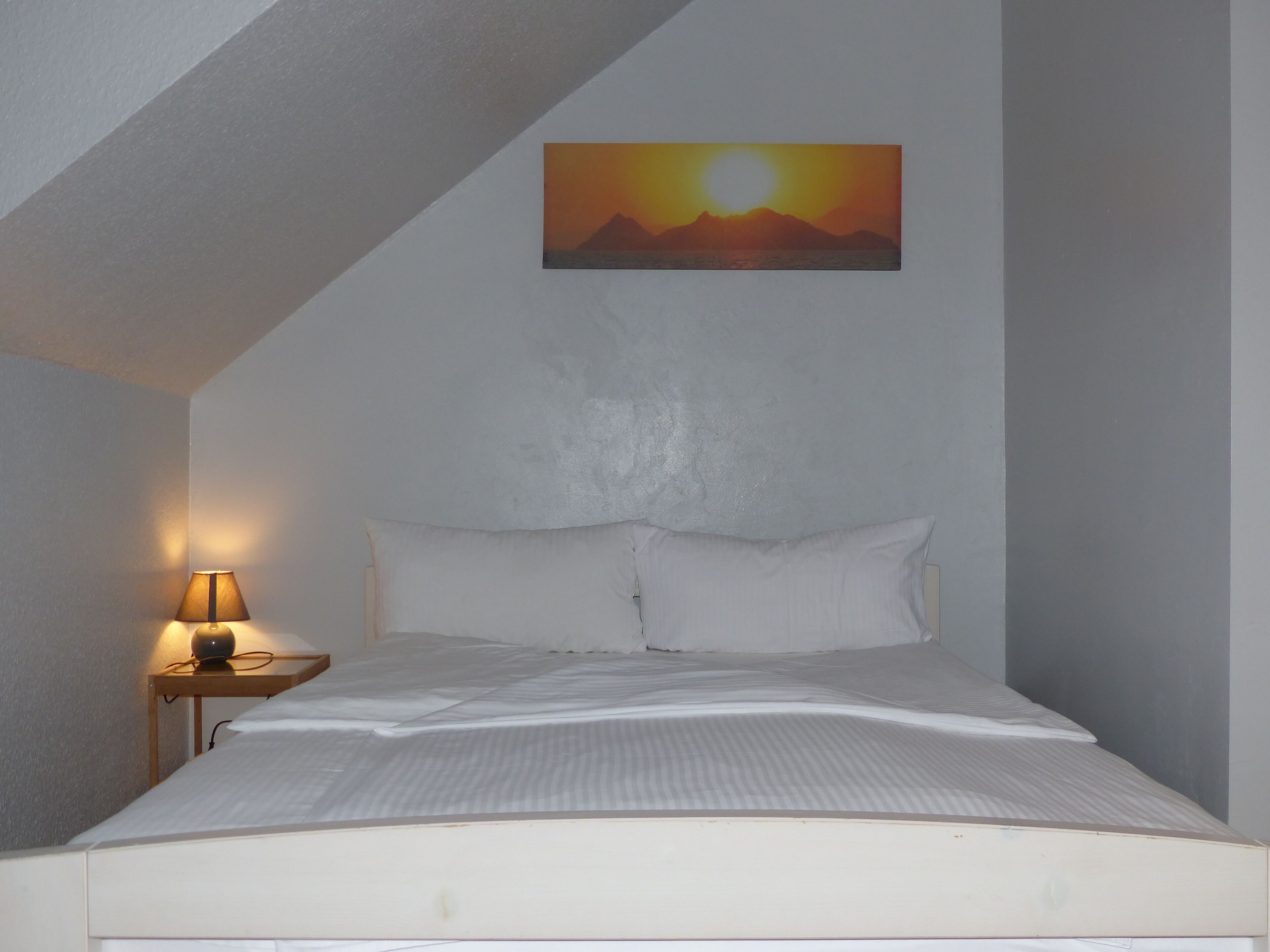 A bedroom with a bed, pillows, a nightstand with a lamp, and a painting of a sunset on the wall.