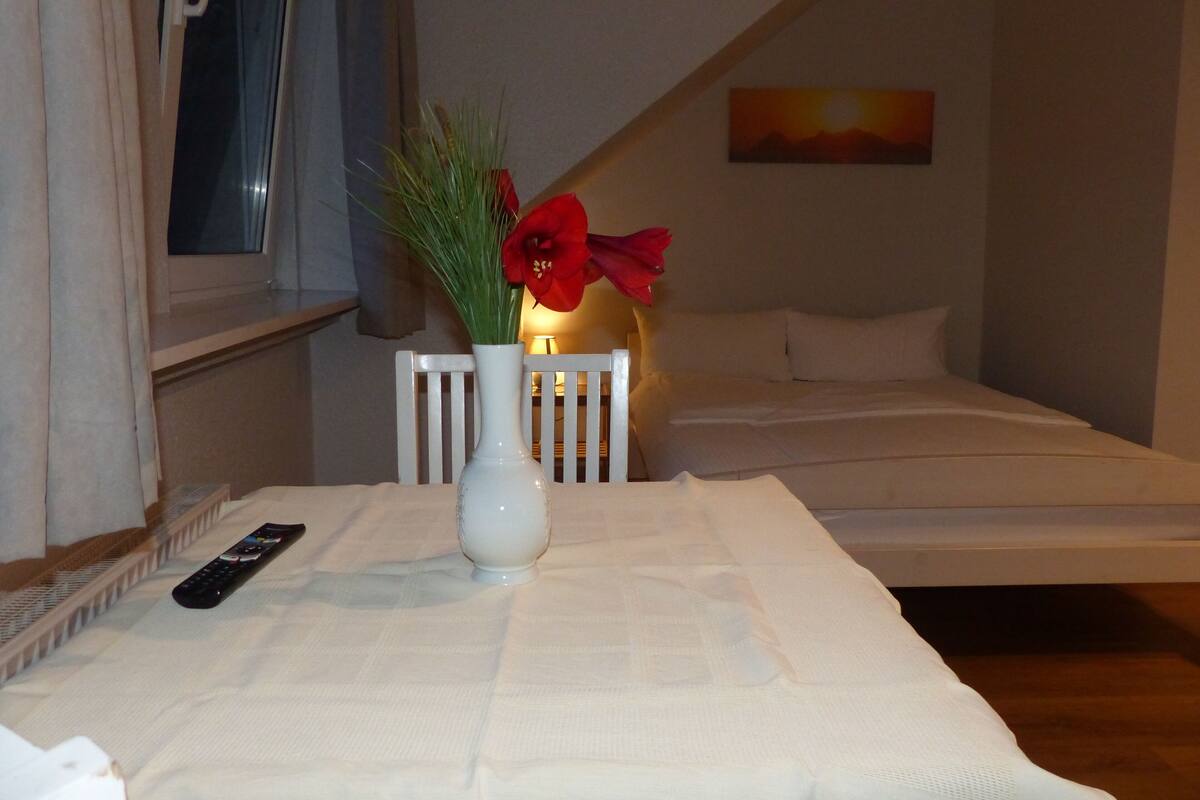 Ein Schlafzimmer mit einem Bett, einem Nachttisch, einer Vase mit Blumen, einer Fernbedienung und einem Gemälde an der Wand.